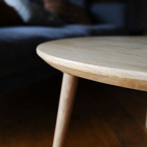 Puede incluir: Una mesa de centro redonda de madera clara con tres patas. La mesa tiene un acabado de veta de madera natural y un borde liso y redondeado. Las patas son delgadas e inclinadas, proporcionando estabilidad.