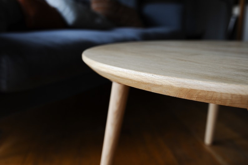 Puede incluir: Una mesa de centro redonda de madera clara con tres patas. La mesa tiene un acabado de veta de madera natural y un borde liso y redondeado. Las patas son delgadas e inclinadas, proporcionando estabilidad.