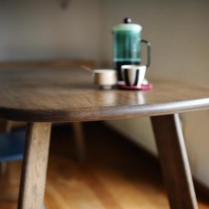 May include: A wooden dining table with a rounded edge and three legs. A French press, a small cup, and a cup with a black and white design are on the table. The table is made of dark wood.