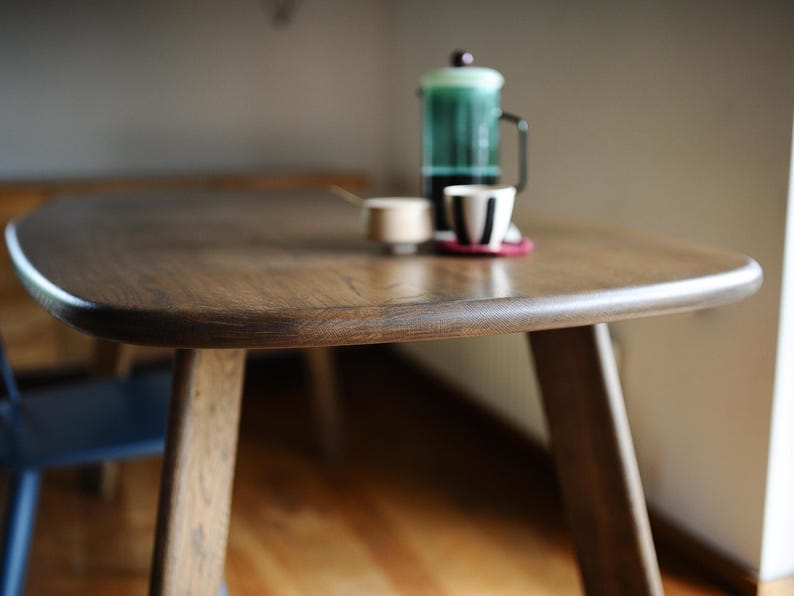 May include: A wooden dining table with a rounded edge and three legs. A French press, a small cup, and a cup with a black and white design are on the table. The table is made of dark wood.