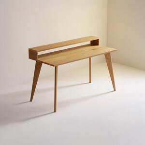 Könnte beinhalten: Ein Schreibtisch aus hellem Holz mit einer rechteckigen, flachen Arbeitsfläche und vier konisch zulaufenden Beinen. Der Schreibtisch verfügt über ein erhöhtes, schmales horizontales Regal an der Hinterkante, das eine abgestufte Struktur bildet.
