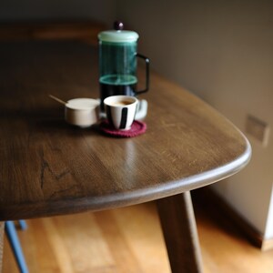 May include: A dark brown wooden table with a rounded edge, holding a French press, a cup of coffee, a small sugar bowl, and a red coaster. The table is supported by wooden legs.