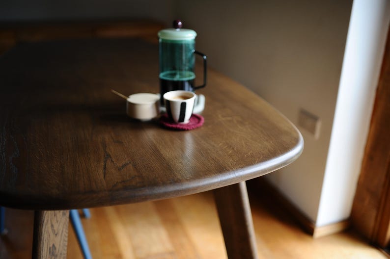 May include: A dark brown wooden table with a rounded edge, holding a French press, a cup of coffee, a small sugar bowl, and a red coaster. The table is supported by wooden legs.