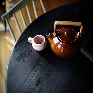 Puede incluir: Una tetera marrón con asa de madera y una taza de té rosa sobre una mesa de madera negra.