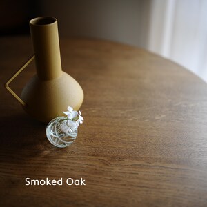 May include: A mustard-colored vase with a handle and a small, clear glass vase holding white flowers. The vases sit on a round, brown wooden table. The text "Smoked Oak" is at the bottom of the image.