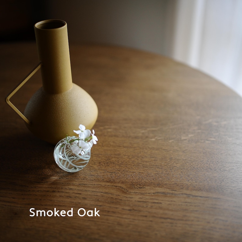 May include: A mustard-colored vase with a handle and a small, clear glass vase holding white flowers. The vases sit on a round, brown wooden table. The text "Smoked Oak" is at the bottom of the image.