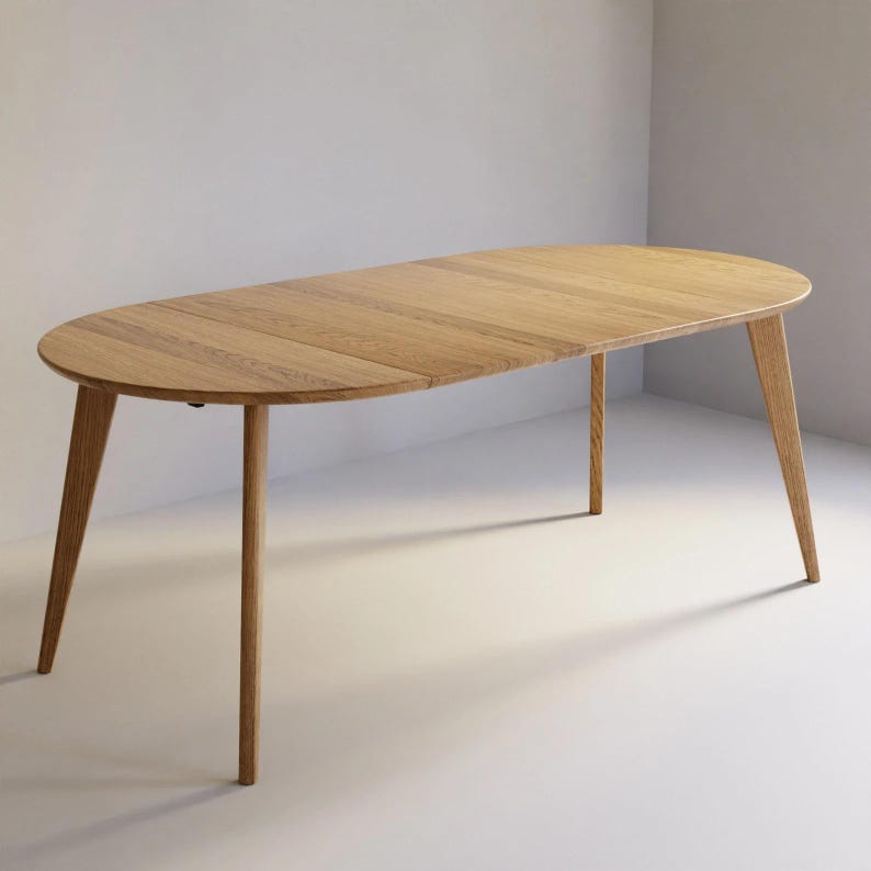 May include: An oval dining table crafted from light-toned wood with a visible grain pattern. The table features four tapered legs that extend outward from the center, supporting a smooth, flat surface with a distinct seam across the middle.
