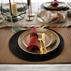 May include: A place setting featuring a speckled beige ceramic plate, a red linen napkin secured with a dark ring, gold-toned flatware, and a small sprig of rosemary, all arranged on a round black woven placemat atop a wooden dining table.