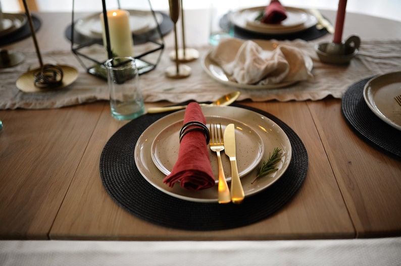 May include: A place setting featuring a speckled beige ceramic plate, a red linen napkin secured with a dark ring, gold-toned flatware, and a small sprig of rosemary, all arranged on a round black woven placemat atop a wooden dining table.