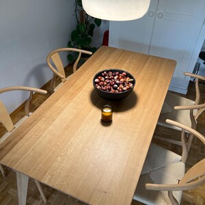 Puede incluir: Una mesa de comedor rectangular de madera clara con cuatro sillas de madera de respaldo curvo y asientos trenzados. Un cuenco negro lleno de casta&ntilde;as marrones y una peque&ntilde;a vela en un frasco de vidrio &aacute;mbar sobre la mesa bajo una l&aacute;mpara.