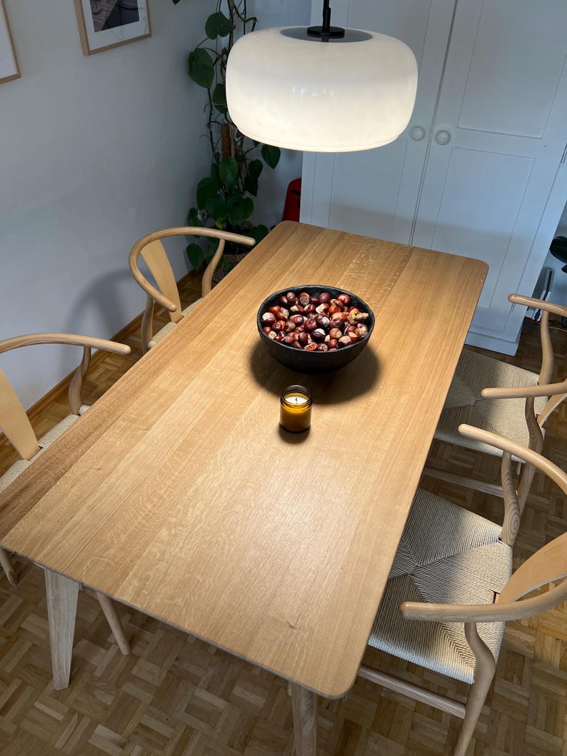 Puede incluir: Una mesa de comedor rectangular de madera clara con cuatro sillas de madera de respaldo curvo y asientos trenzados. Un cuenco negro lleno de casta&ntilde;as marrones y una peque&ntilde;a vela en un frasco de vidrio &aacute;mbar sobre la mesa bajo una l&aacute;mpara.