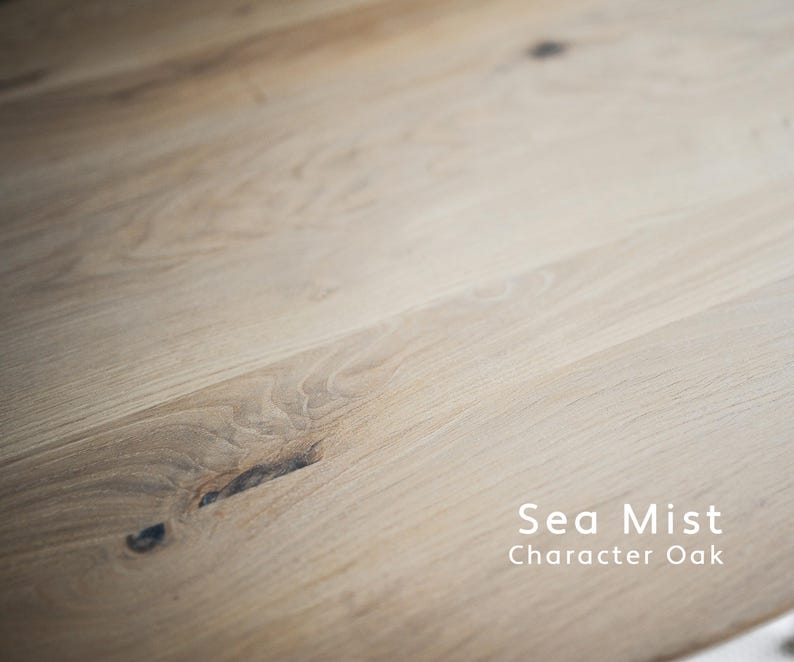 Puede incluir: Una superficie de madera de tono claro con un patr&oacute;n de veta visible y nudos naturales, que presenta el texto Sea Mist Character Oak en la esquina inferior derecha. La madera muestra un acabado liso con sutiles variaciones de color.