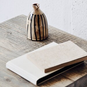 May include: A small, brown ceramic vase with black vertical stripes sits on a wooden table. Two books, one white and one brown, are stacked on the table.