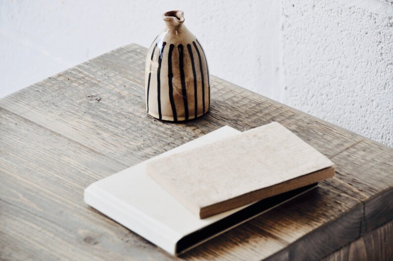 May include: A small, brown ceramic vase with black vertical stripes sits on a wooden table. Two books, one white and one brown, are stacked on the table.