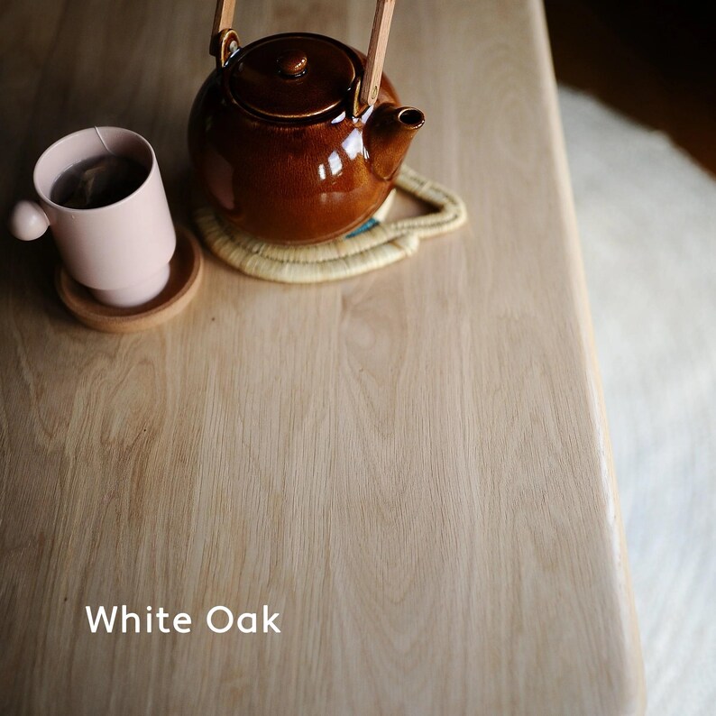 May include: A brown ceramic teapot with a wooden handle sits on a woven coaster next to a pink mug with tea. The objects are on a light-colored wooden surface. The text "White Oak" is at the bottom.