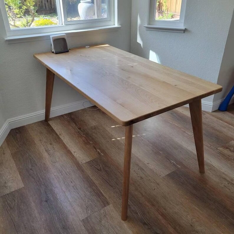 Puede incluir: Una mesa de comedor rectangular de madera clara que presenta una superficie lisa y plana con patrones de veta de madera natural visibles y cuatro patas c&oacute;nicas e inclinadas que se extienden hacia afuera desde las esquinas del mueble.