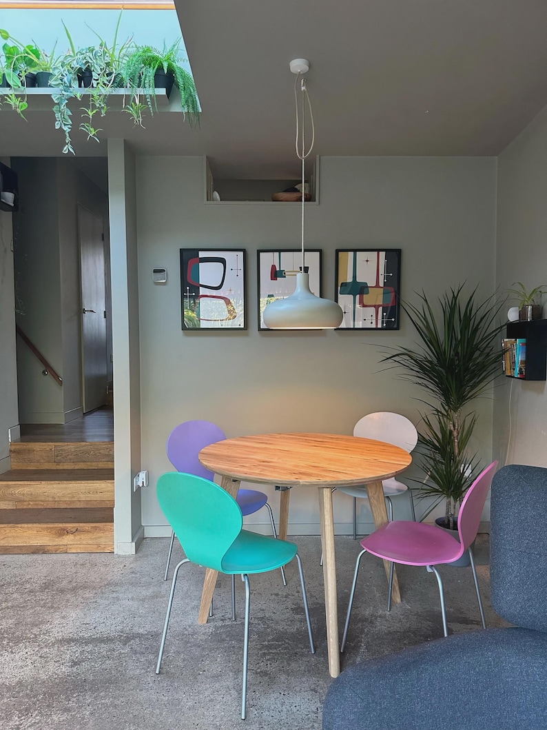 May include: A round wooden dining table with four chairs in purple, teal, white, and pink, each featuring thin metal legs. A light gray pendant lamp hangs above the table, and three framed abstract art prints are mounted on the gray wall.
