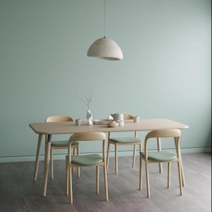 Op de afbeelding: Een eettafel van licht hout met vier bijpassende stoelen. De stoelen hebben licht houten frames en groen gestoffeerde zittingen. Boven de tafel, die is gedekt met een vaas, kopjes en een theepot, hangt een witte hanglamp.