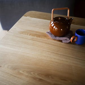 Puede incluir: Una tetera de cer&aacute;mica marr&oacute;n brillante con un asa de madera clara colocada sobre una peque&ntilde;a servilleta de tela plegada con rayas granates y blancas junto a una taza de cer&aacute;mica azul s&oacute;lida sobre una superficie de madera clara con vetas.