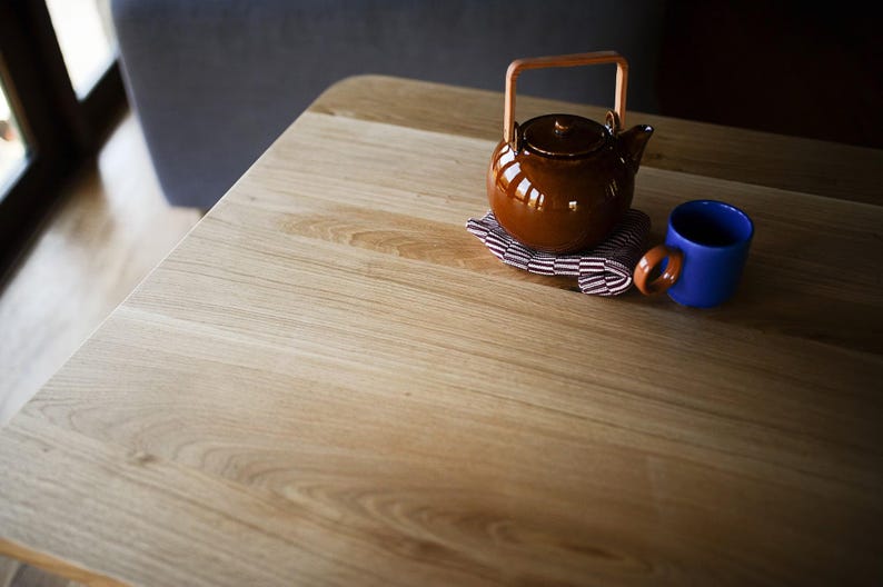 Puede incluir: Una tetera de cer&aacute;mica marr&oacute;n brillante con un asa de madera clara colocada sobre una peque&ntilde;a servilleta de tela plegada con rayas granates y blancas junto a una taza de cer&aacute;mica azul s&oacute;lida sobre una superficie de madera clara con vetas.