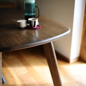 May include: A wooden table with a dark finish, holding a French press, a small cup with a black and white design, and a small sugar bowl. The table has a rounded edge and is supported by angled legs. A red coaster sits under the cup.