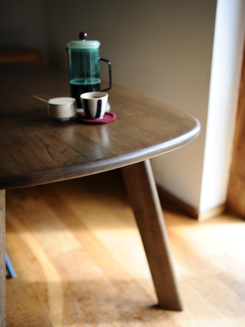 May include: A wooden table with a dark finish, holding a French press, a small cup with a black and white design, and a small sugar bowl. The table has a rounded edge and is supported by angled legs. A red coaster sits under the cup.