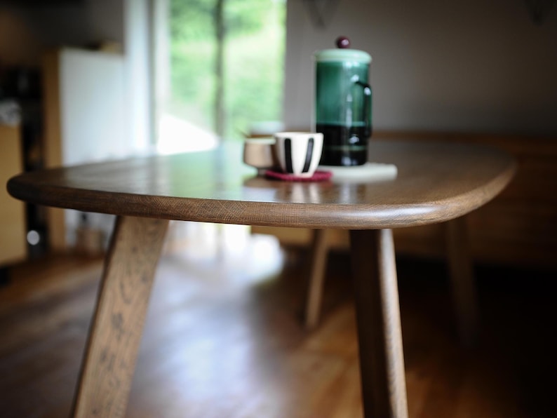 May include: A wooden, oval-shaped coffee table with a dark finish. The table has four angled legs and a smooth surface. On the table are a small cup with black stripes, a small white cup, and a green glass carafe with a red top.