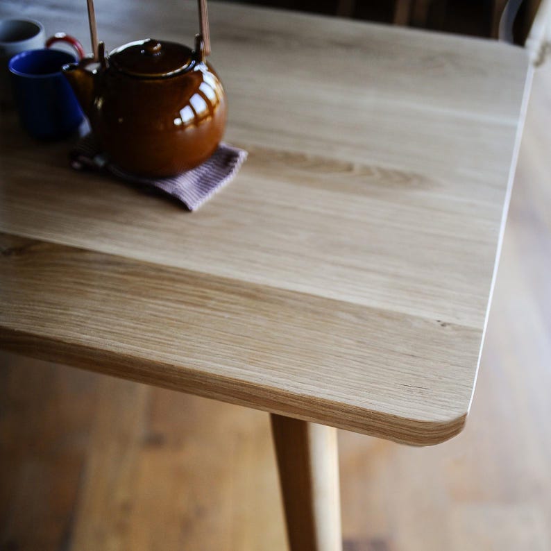 Puede incluir: Una mesa de madera clara con una superficie rectangular lisa y esquinas redondeadas. Una tetera de cer&aacute;mica marr&oacute;n brillante con asa de metal descansa sobre un peque&ntilde;o posavasos de tela texturizada, acompa&ntilde;ada por una taza azul y una taza blanca.