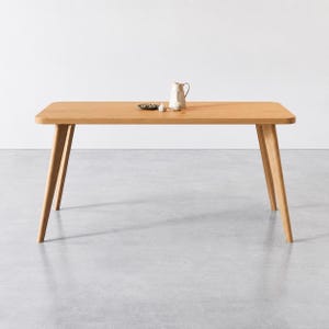 Puede incluir: Una mesa de comedor rectangular de madera clara con esquinas redondeadas y cuatro patas inclinadas. Sobre la superficie de la mesa hay una peque&ntilde;a jarra de cer&aacute;mica blanca, un diente de ajo y un plato peque&ntilde;o oscuro con objetos verdes.