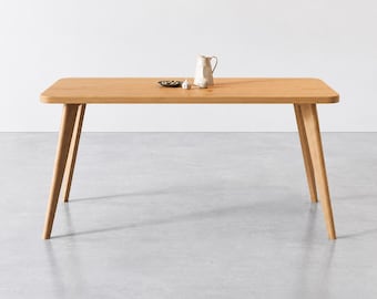 Op de afbeelding: Een rechthoekige eettafel van lichtgekleurd hout met afgeronde hoeken en vier schuine poten. Op het tafeloppervlak staan een kleine witte keramische kan, een bol knoflook en een klein donker schaaltje met enkele kleine groene voorwerpen.