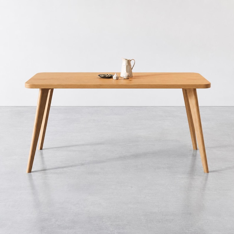 Puede incluir: Una mesa de comedor rectangular de madera clara con esquinas redondeadas y cuatro patas inclinadas. Sobre la superficie de la mesa hay una peque&ntilde;a jarra de cer&aacute;mica blanca, un diente de ajo y un plato peque&ntilde;o oscuro con objetos verdes.