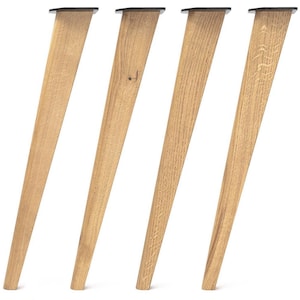 May include: A set of four tapered furniture legs crafted from light-toned wood with a visible natural grain pattern. Each leg features a flat, black metal mounting plate at the top, angled to provide a slanted orientation for furniture support.