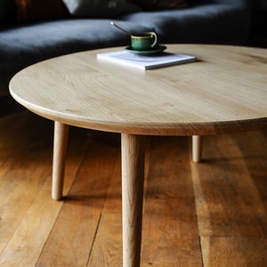 Puede incluir: Una mesa de centro redonda de madera clara con cuatro patas c&oacute;nicas. Una taza y platillo verdes con una cuchara descansan sobre un libro. La mesa est&aacute; sobre un suelo de madera.