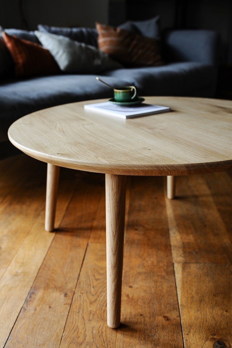 Puede incluir: Una mesa de centro redonda de madera clara con cuatro patas c&oacute;nicas. Una taza y platillo verdes con una cuchara descansan sobre un libro. La mesa est&aacute; sobre un suelo de madera.