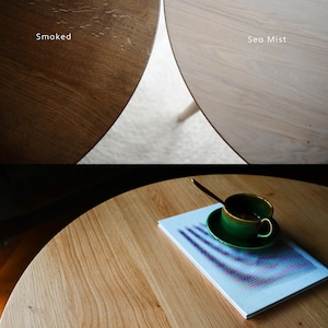 May include: A round wooden table surface shown in two finishes: a dark brown labeled Smoked and a light beige labeled Sea Mist. The bottom section displays a light wood table with a green ceramic cup, a saucer, and a book with a blue graphic cover.