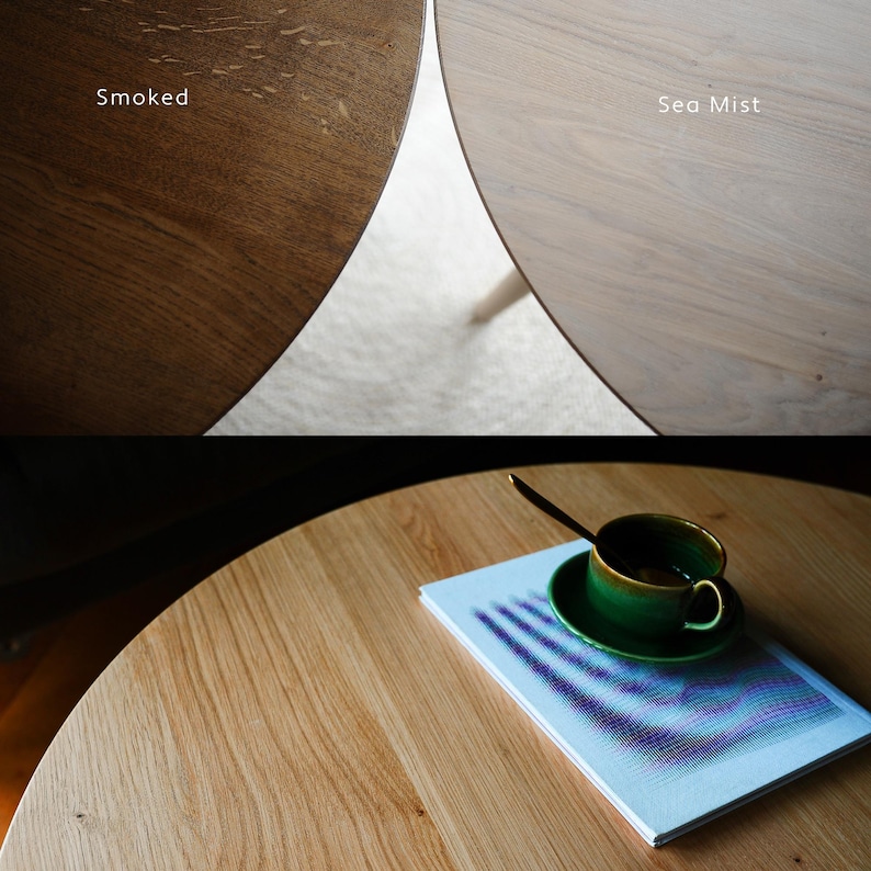 May include: A round wooden table surface shown in two finishes: a dark brown labeled Smoked and a light beige labeled Sea Mist. The bottom section displays a light wood table with a green ceramic cup, a saucer, and a book with a blue graphic cover.