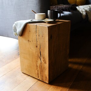 May include: A rustic wooden cube stool with a natural wood finish. The stool is sitting on a hardwood floor and has a white ceramic dish and a black mug on top.