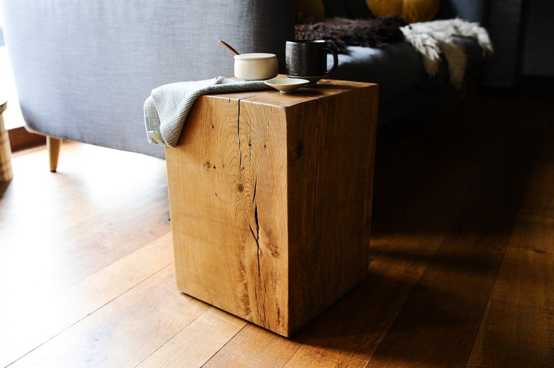 May include: A rustic wooden cube stool with a natural wood finish. The stool is sitting on a hardwood floor and has a white ceramic dish and a black mug on top.