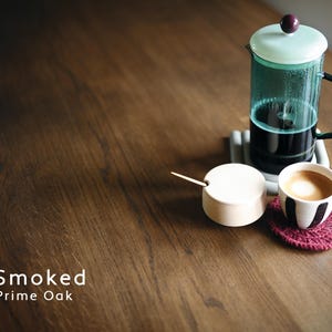 Puede incluir: Una cafetera de &eacute;mbolo con tapa verde claro y asa negra sobre un posavasos gris. Un azucarero blanco peque&ntilde;o con cuchara de madera y una taza de caf&eacute; con dise&ntilde;o blanco y negro sobre mesa de madera. Un posavasos rojo bajo la taza. El texto "Smoked Prime Oak" est&aacute; en la parte inferior.