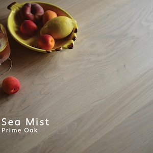 May include: A light-toned oak wood surface with a visible grain pattern, featuring the text Sea Mist Prime Oak. A yellow ceramic bowl shaped like a banana holds various fruits, including a lemon and peaches, next to a glass of iced tea.