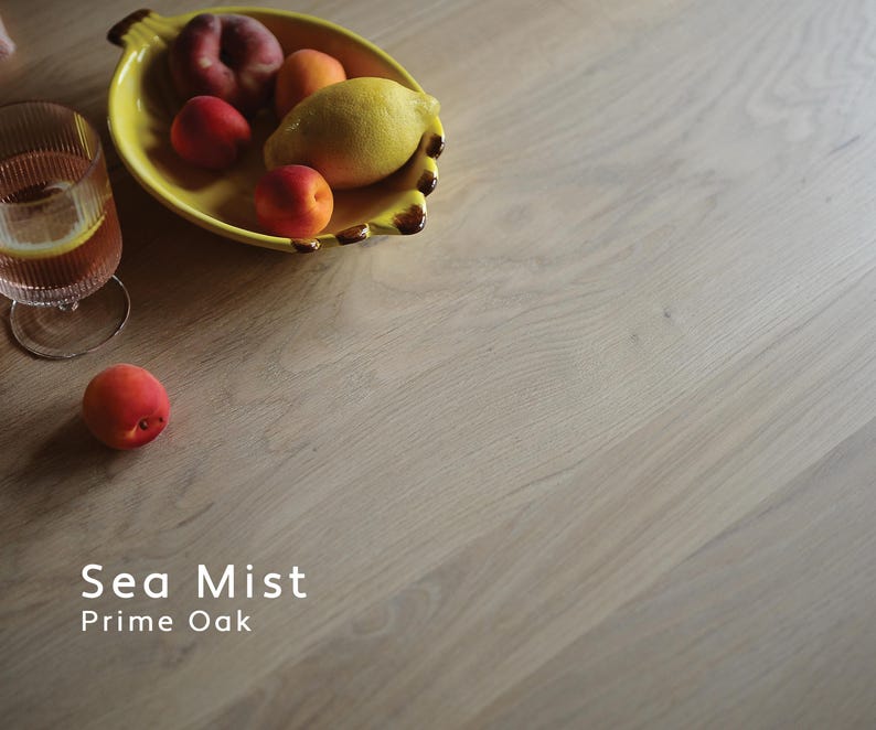 May include: A light-toned oak wood surface with a visible grain pattern, featuring the text Sea Mist Prime Oak. A yellow ceramic bowl shaped like a banana holds various fruits, including a lemon and peaches, next to a glass of iced tea.