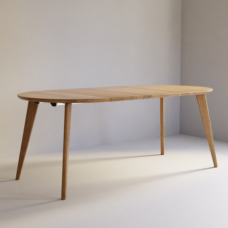 May include: An oval dining table crafted from light-toned wood with a smooth, flat surface and four tapered legs that angle outward. The table features a visible seam across the center, indicating a modular or extendable design construction.