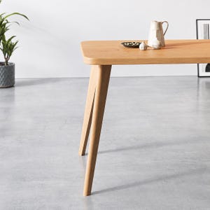 Puede incluir: Una mesa de comedor de madera de color marr&oacute;n claro con esquinas redondeadas y patas inclinadas. Sobre la superficie de la mesa hay un peque&ntilde;o plato negro con aceitunas verdes, una jarra de cer&aacute;mica blanca y un peque&ntilde;o objeto decorativo con forma de ajo.