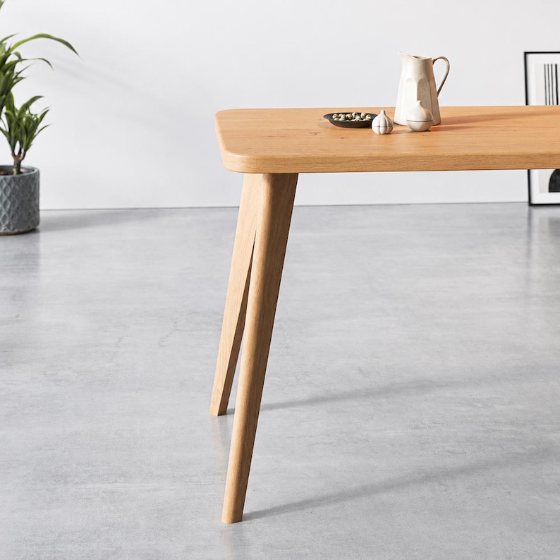 Puede incluir: Una mesa de comedor de madera de color marr&oacute;n claro con esquinas redondeadas y patas inclinadas. Sobre la superficie de la mesa hay un peque&ntilde;o plato negro con aceitunas verdes, una jarra de cer&aacute;mica blanca y un peque&ntilde;o objeto decorativo con forma de ajo.