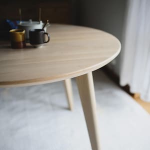 May include: A round light-colored wooden dining table with tapered legs. On the surface of the table, there is a white teapot, a small yellow ceramic cup, and a dark brown ceramic mug. The table has a smooth, natural wood grain finish.