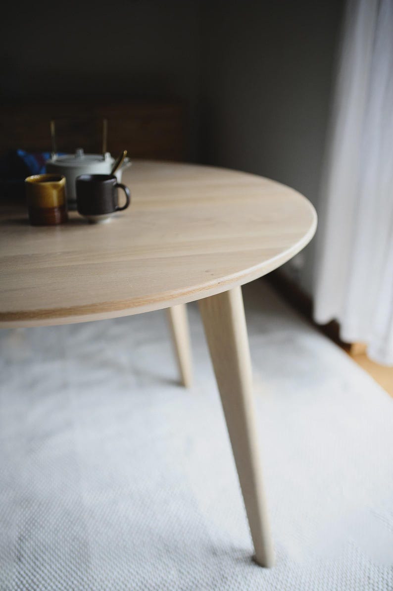 May include: A round light-colored wooden dining table with tapered legs. On the surface of the table, there is a white teapot, a small yellow ceramic cup, and a dark brown ceramic mug. The table has a smooth, natural wood grain finish.