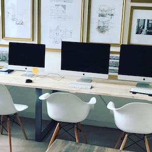 Op de afbeelding: Een lange houten tafel met vier witte stoelen en vier Apple iMac-computers. De computers zijn allemaal aan en hebben zwarte schermen. Er hangen ingelijste foto's aan de muur achter de tafel.