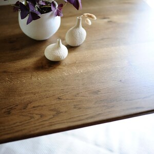 Puede incluir: Una mesa rectangular de madera con veta visible, que presenta una maceta blanca redondeada con una planta de hojas moradas y dos peque&ntilde;os jarrones de cer&aacute;mica blanca texturizada con cuellos estrechos sobre la superficie.
