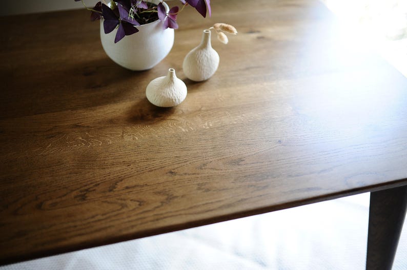 Puede incluir: Una mesa rectangular de madera con veta visible, que presenta una maceta blanca redondeada con una planta de hojas moradas y dos peque&ntilde;os jarrones de cer&aacute;mica blanca texturizada con cuellos estrechos sobre la superficie.