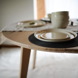 May include: A round light-colored wooden dining table featuring a black circular placemat, a set of stacked cream-colored ceramic plates, and two clear glass tumblers. A light-colored table runner and additional stacked bowls are visible.